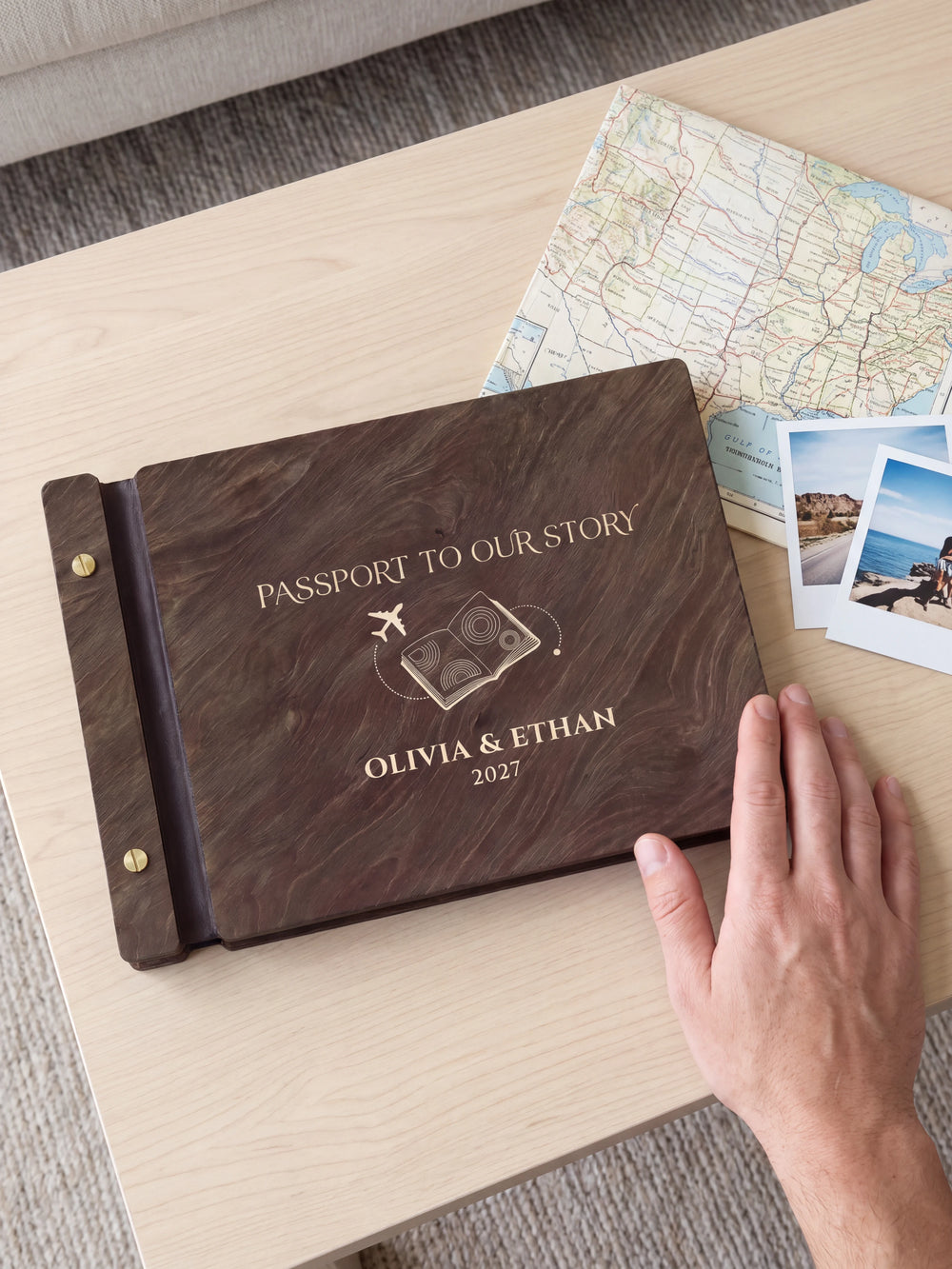 A hand rests beside the closed dark brown Jacobean album on a table. Cream engraving and personalized wording are visible, with travel photos nearby.
