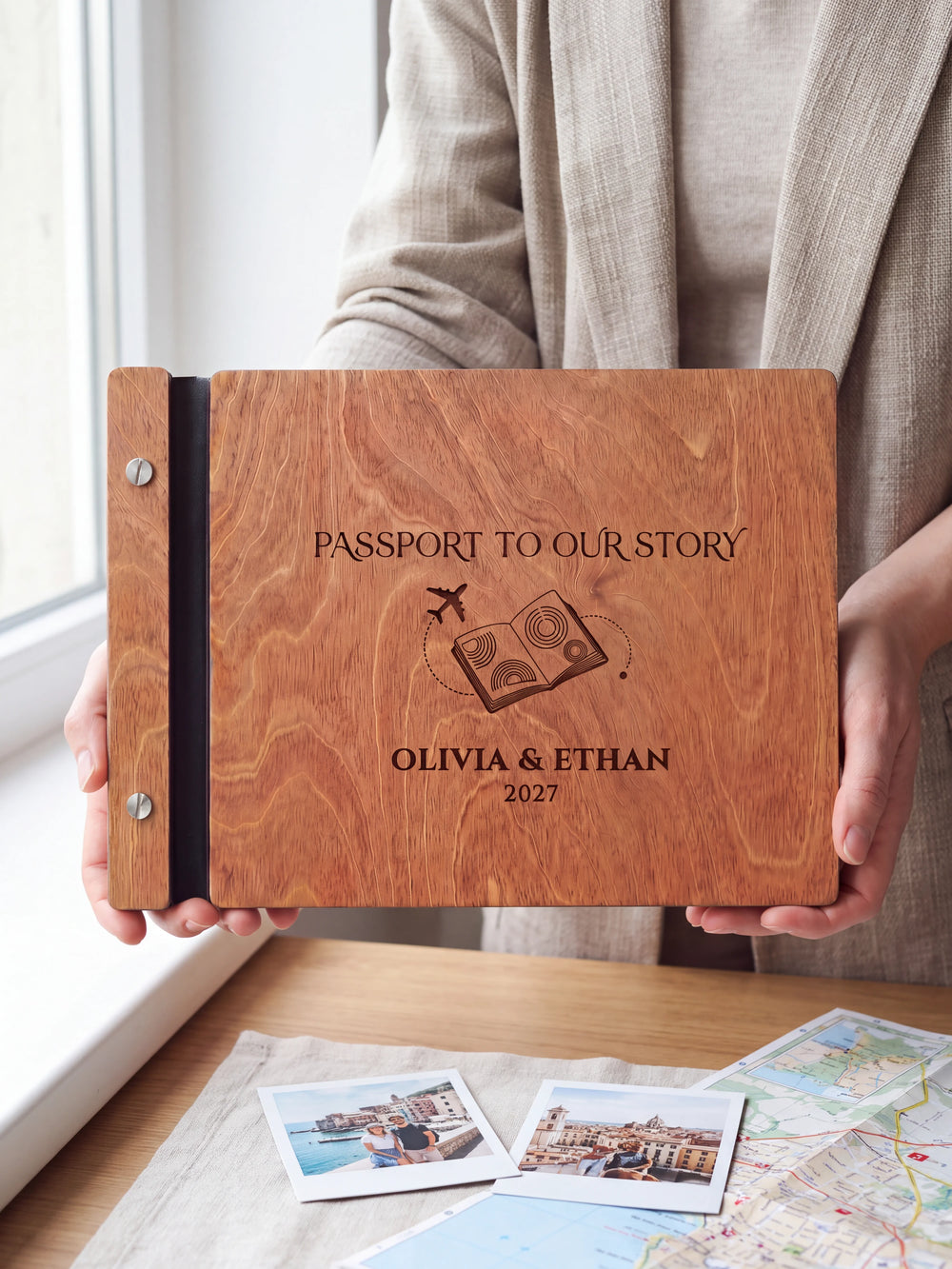 Hands hold the Hazelnut wooden album near a window. The full cover displays a passport and airplane in dark engraving, with personalized lettering and a black binding strip.