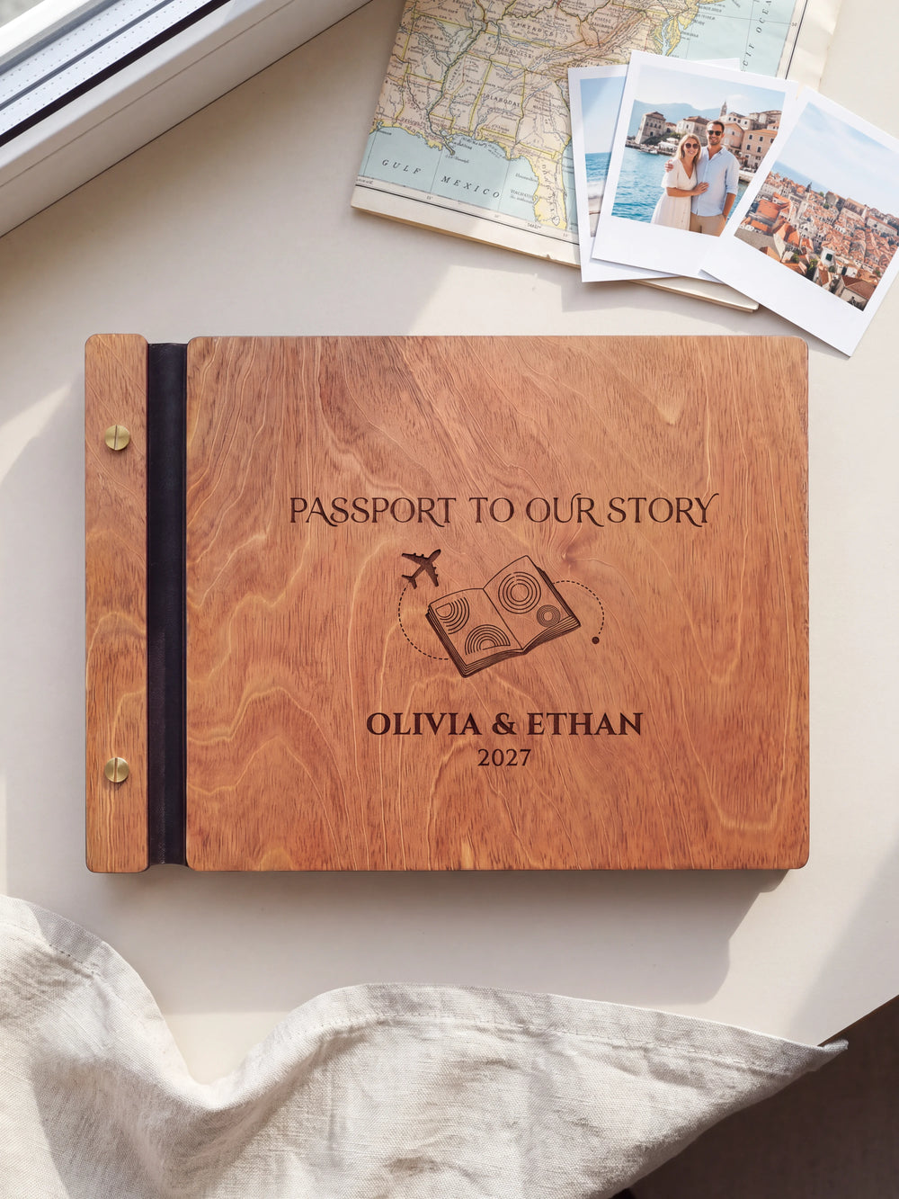 Hazelnut wooden album on a light tabletop with neutral linen. The full cover shows a passport and airplane, personalized wording and the two-screw binding.