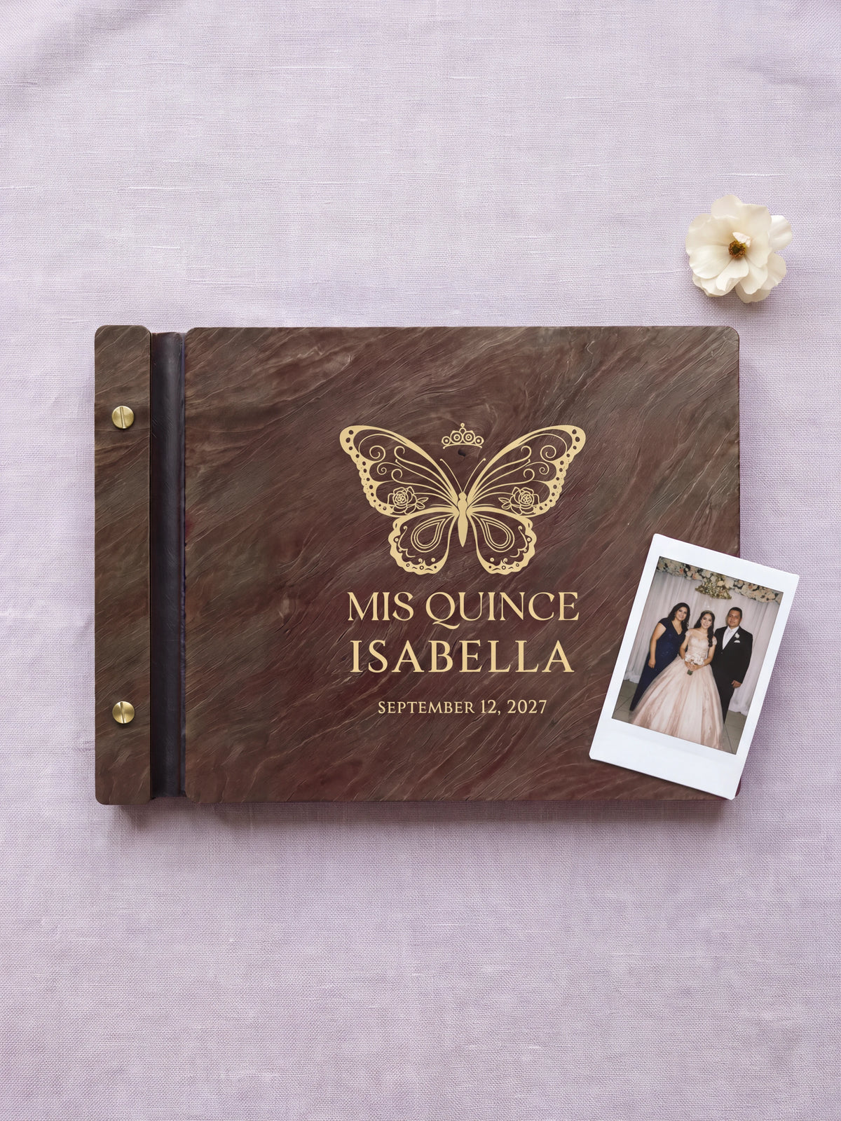 Jacobean wood quinceañera photo guest book lying flat on pale lilac linen, the cover engraved with “MIS QUINCE”, “ISABELLA” and “SEPTEMBER 12, 2027”; two gold-tone screws along the black vegan leather spine