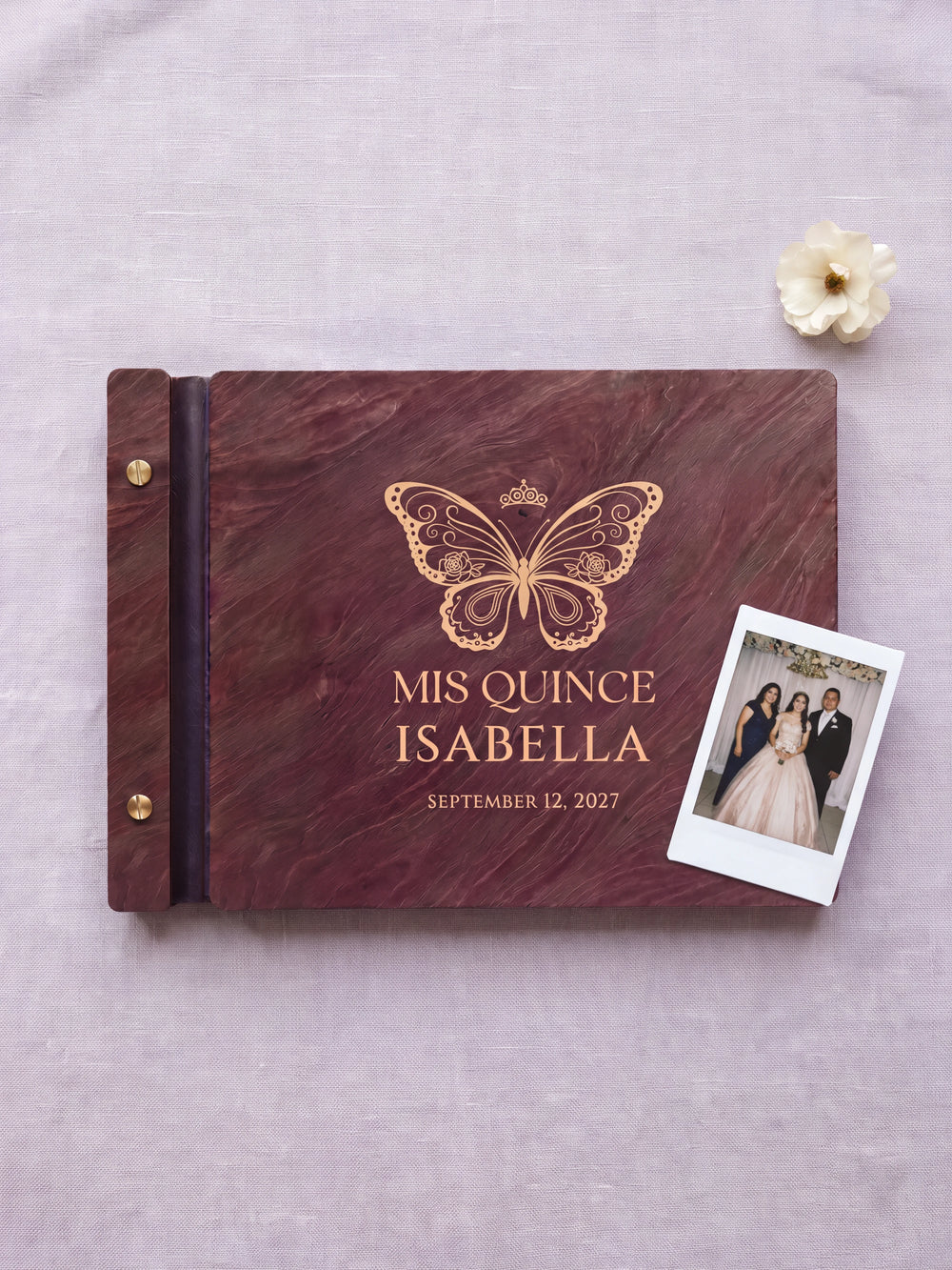 Jacobean wood quinceañera photo guest book lying flat on pale lilac linen, the cover engraved with “MIS QUINCE”, “ISABELLA” and “SEPTEMBER 12, 2027”; two gold-tone screws along the black vegan leather spine