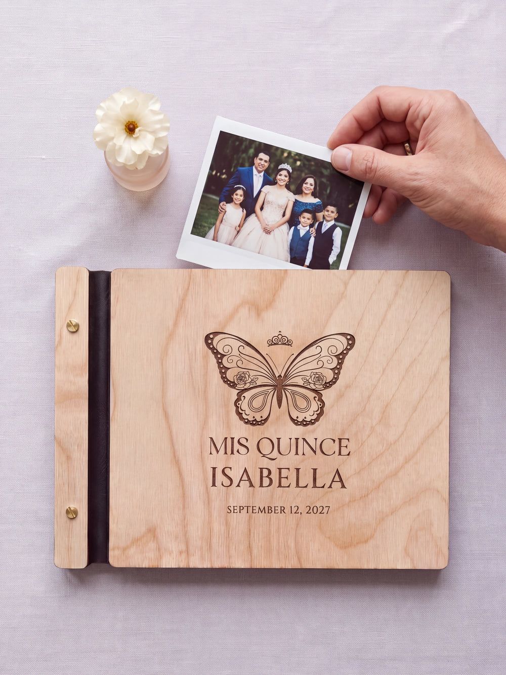 Natural wood quinceañera photo guest book on a light table while a hand picks an instant photo to add; the engraved cover reads “MIS QUINCE”, “ISABELLA” and “SEPTEMBER 12, 2027”