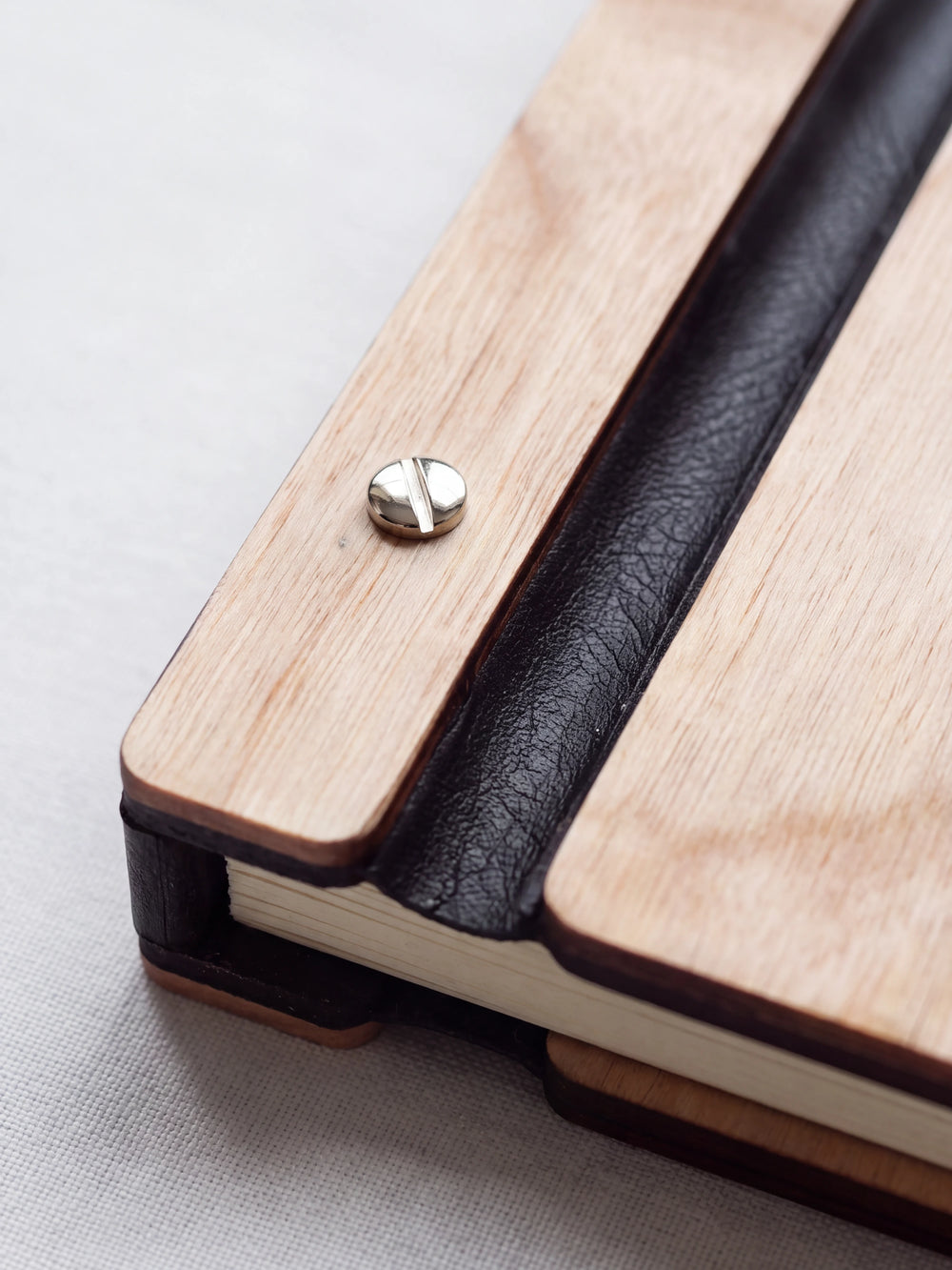 Close-up of a polished silver screw in the Natural wood strip beside the black vegan leather binding, the cream sheet stack visible below