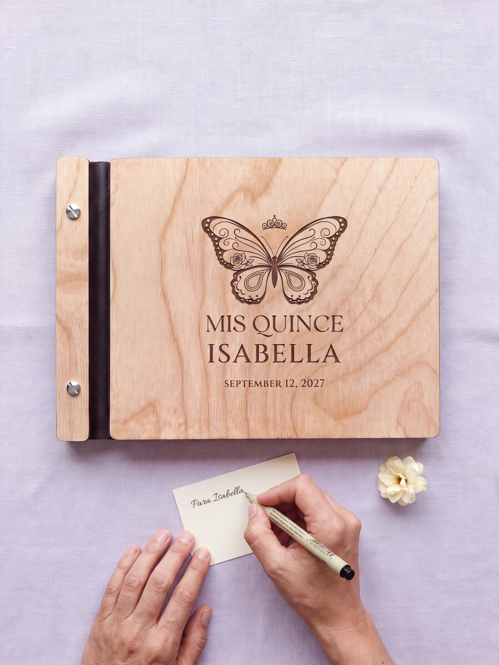 A hand writing a small gift card beside the closed Natural wood quinceañera photo guest book; the engraved cover and silver screws are visible