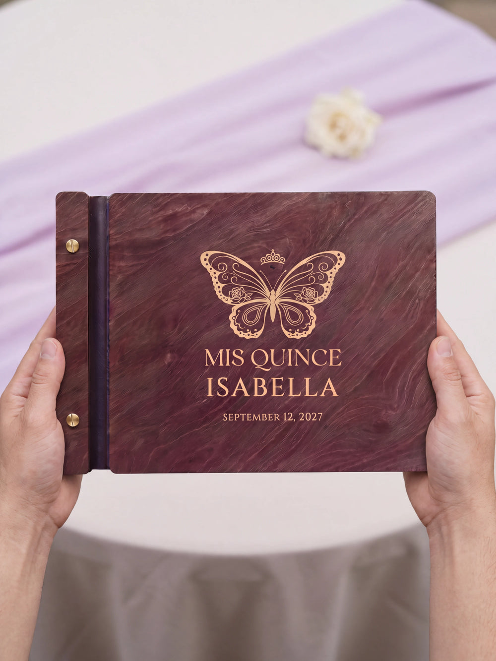 Two hands presenting the closed Jacobean quinceañera photo guest book with its cream engraving of an ornate butterfly topped with a small crown, a small ivory flower beside them