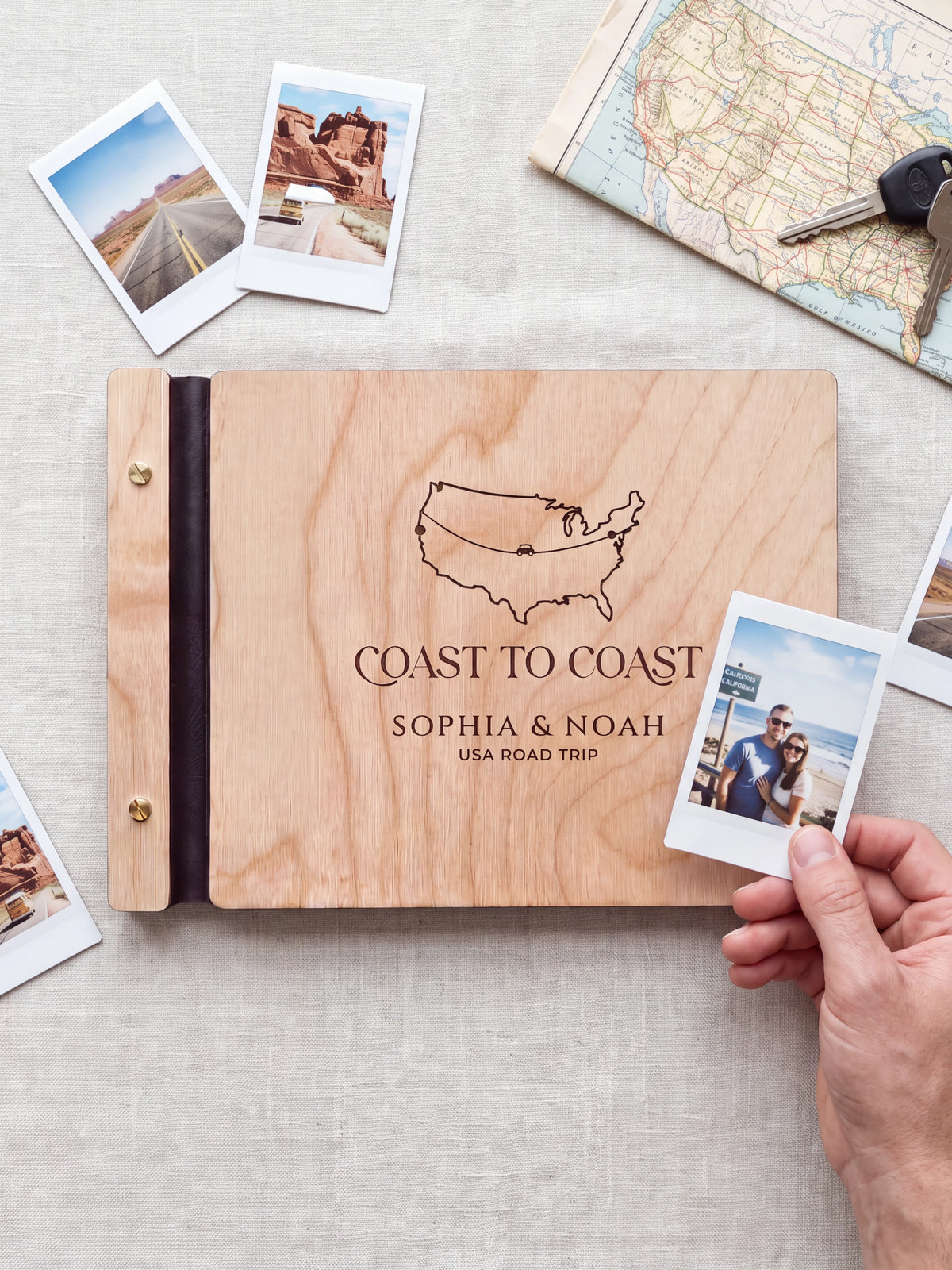 Natural wooden album with dark engraving of a USA outline map and road-trip route. A hand holds an instant photo beside the closed cover, with loose snapshots nearby.
