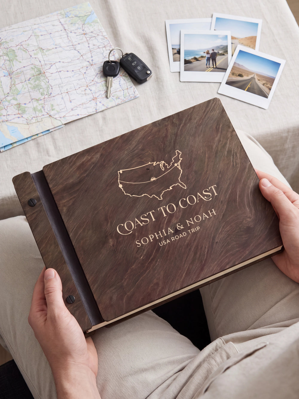 Hands hold a dark brown Jacobean wooden album close to the camera. Cream engraving shows a USA outline map and road-trip route, personalized wording and the black binding strip.