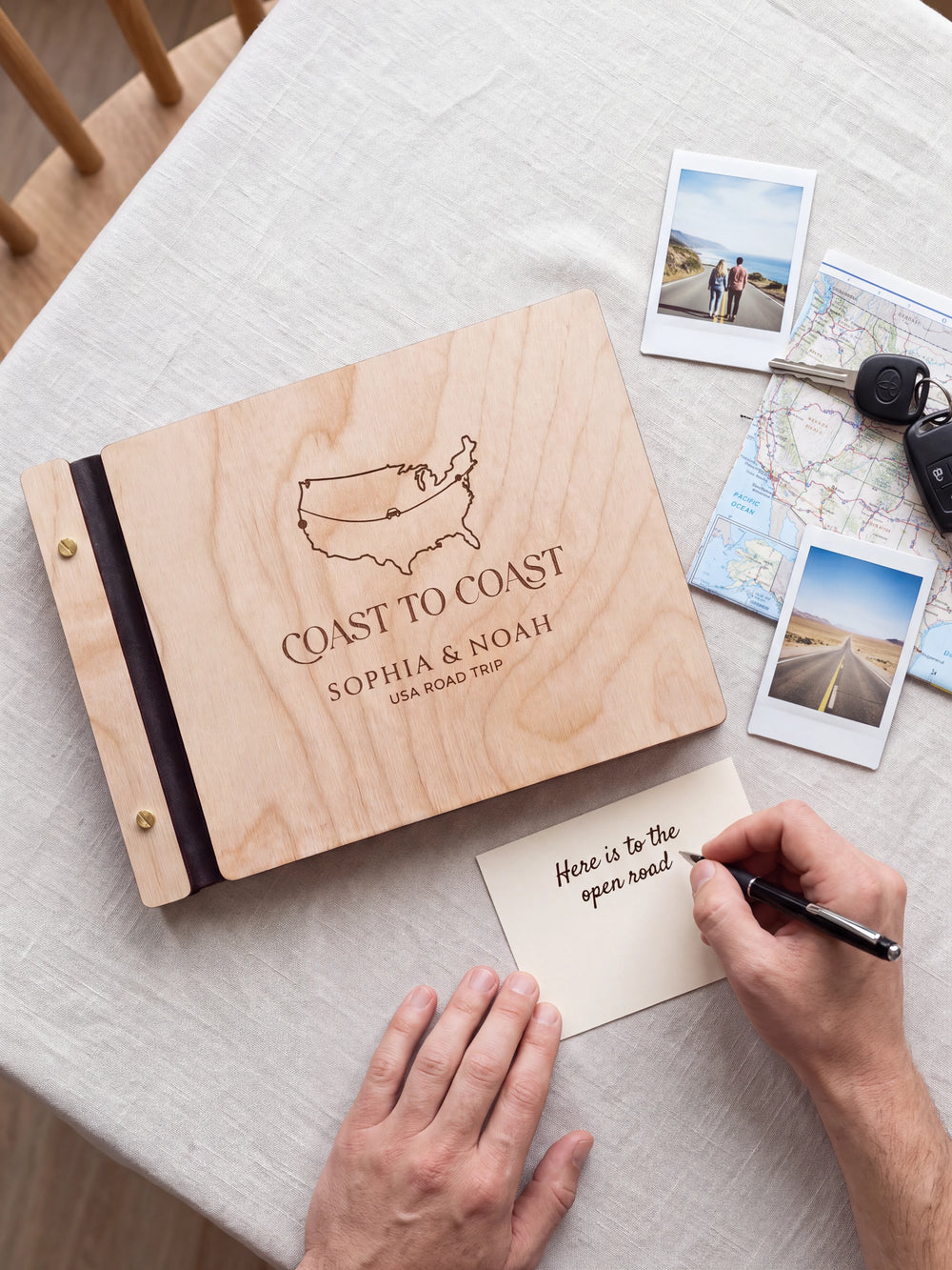 A Natural wooden album showing a USA outline map and road-trip route rests beside a separate gift card. A hand writes 