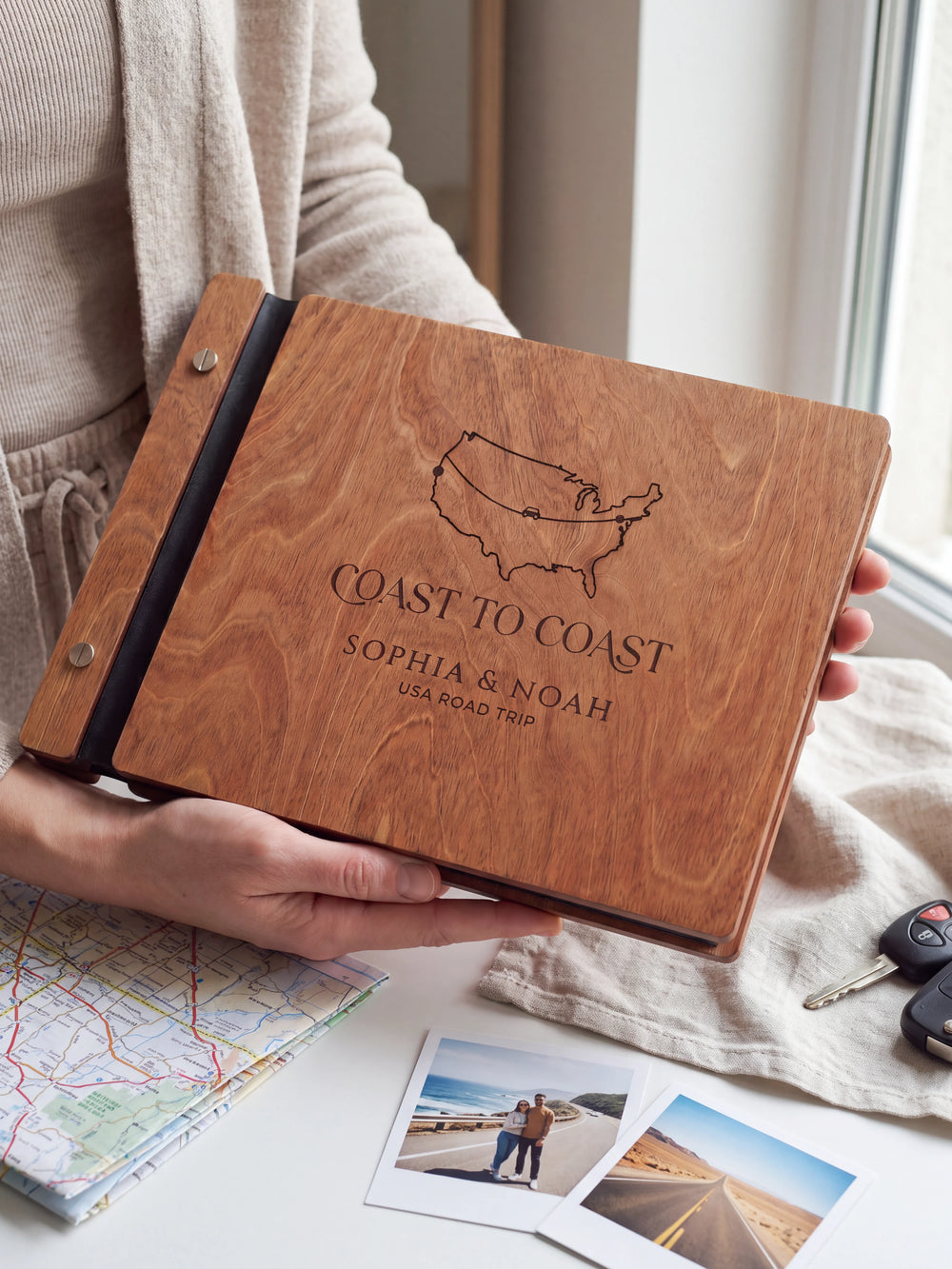 Hands hold the Hazelnut wooden album near a window. The full cover displays a USA outline map and road-trip route in dark engraving, with personalized lettering and a black binding strip.