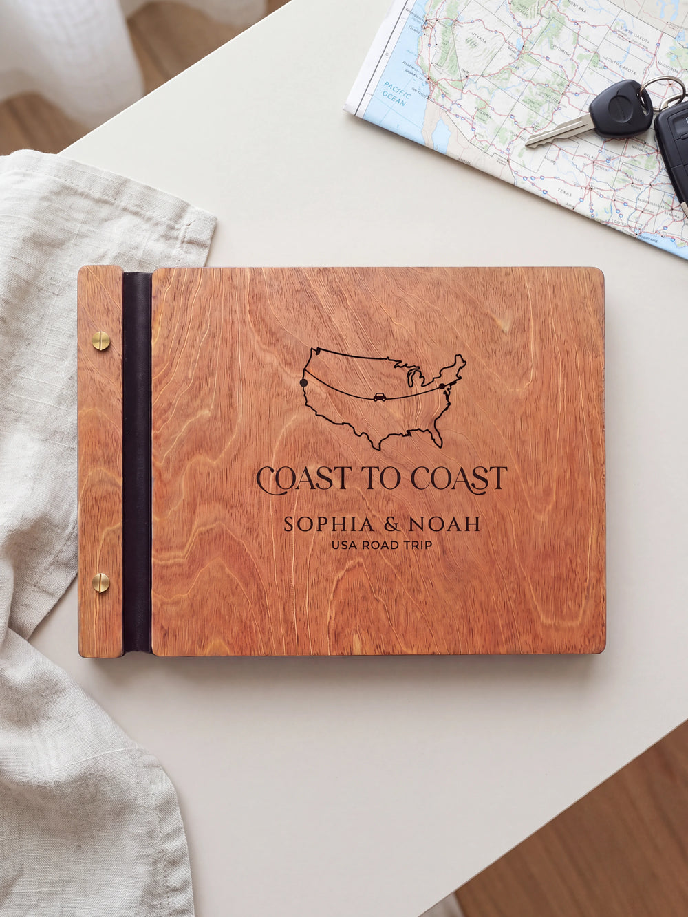 Hazelnut wooden album on a light tabletop with neutral linen. The full cover shows a USA outline map and road-trip route, personalized wording and the two-screw binding.