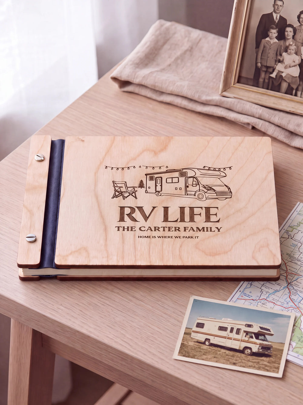 Natural wooden album with brown laser engraving of motorhome, camp chair and string lights, shown in a close lifestyle view with navy blue binding and silver screws. The cover reads RV LIFE.