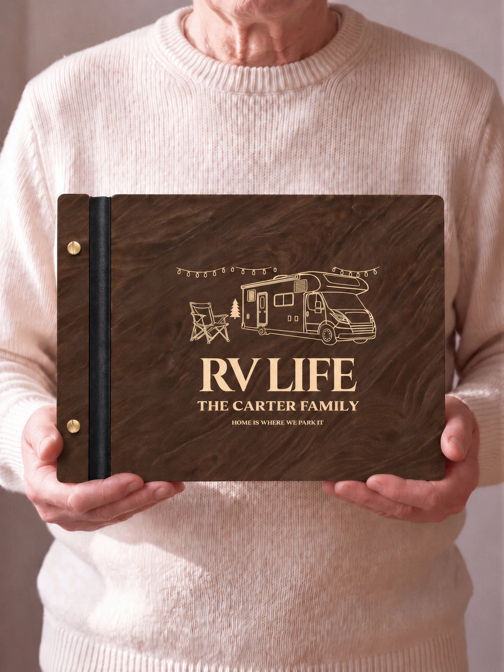 Two hands hold a Jacobean wooden photo album with the RV LIFE cover facing the camera. Cream engraved artwork, the narrow binding and gold screws are visible.
