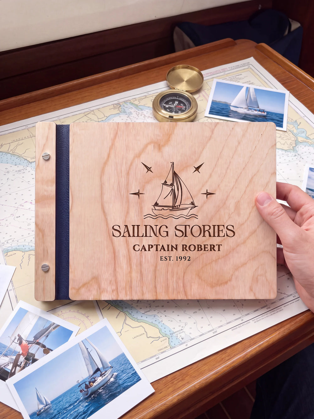 Natural wooden album with brown laser engraving of sailboat, waves and compass stars, shown in a close lifestyle view with navy blue binding and silver screws. The cover reads SAILING STORIES.