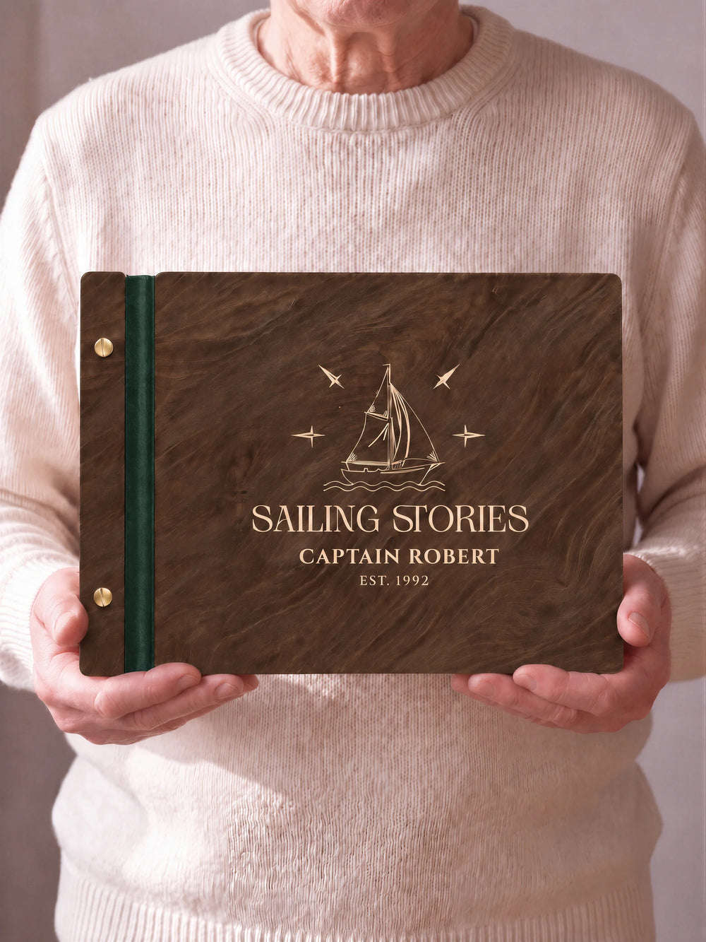 Two hands hold a Jacobean wooden photo album with the SAILING STORIES cover facing the camera. Cream engraved artwork, the narrow binding and gold screws are visible.