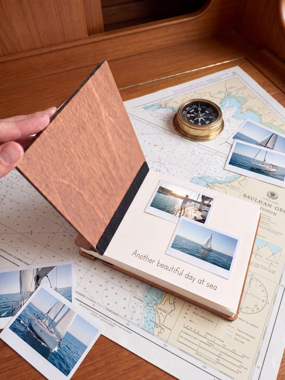 A hand lifts the Hazelnut album’s front cover, revealing a flat black inner fabric strip and a cream page arranged with instant photographs and a handwritten memory.