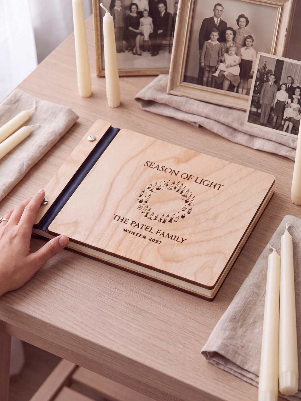 Natural wooden album with brown laser engraving of wreath formed by varied candles, shown in a close lifestyle view with navy blue binding and silver screws. The cover reads SEASON OF LIGHT.