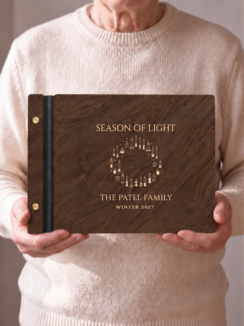 Two hands hold a Jacobean wooden photo album with the SEASON OF LIGHT cover facing the camera. Cream engraved artwork, the narrow binding and gold screws are visible.