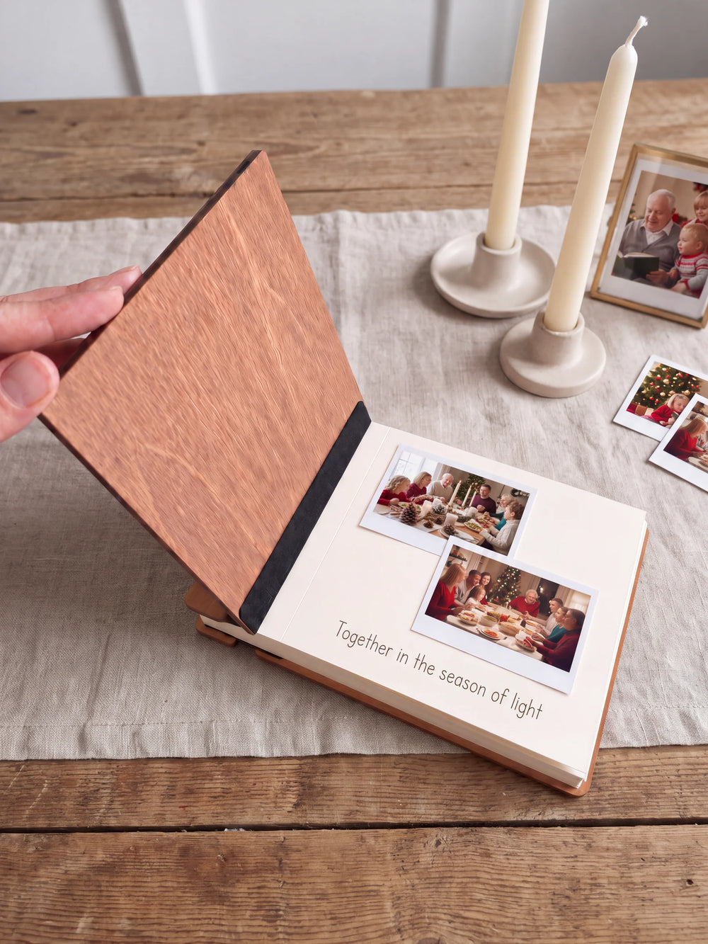 A hand lifts the Hazelnut album’s front cover, revealing a flat black inner fabric strip and a cream page arranged with instant photographs and a handwritten memory.