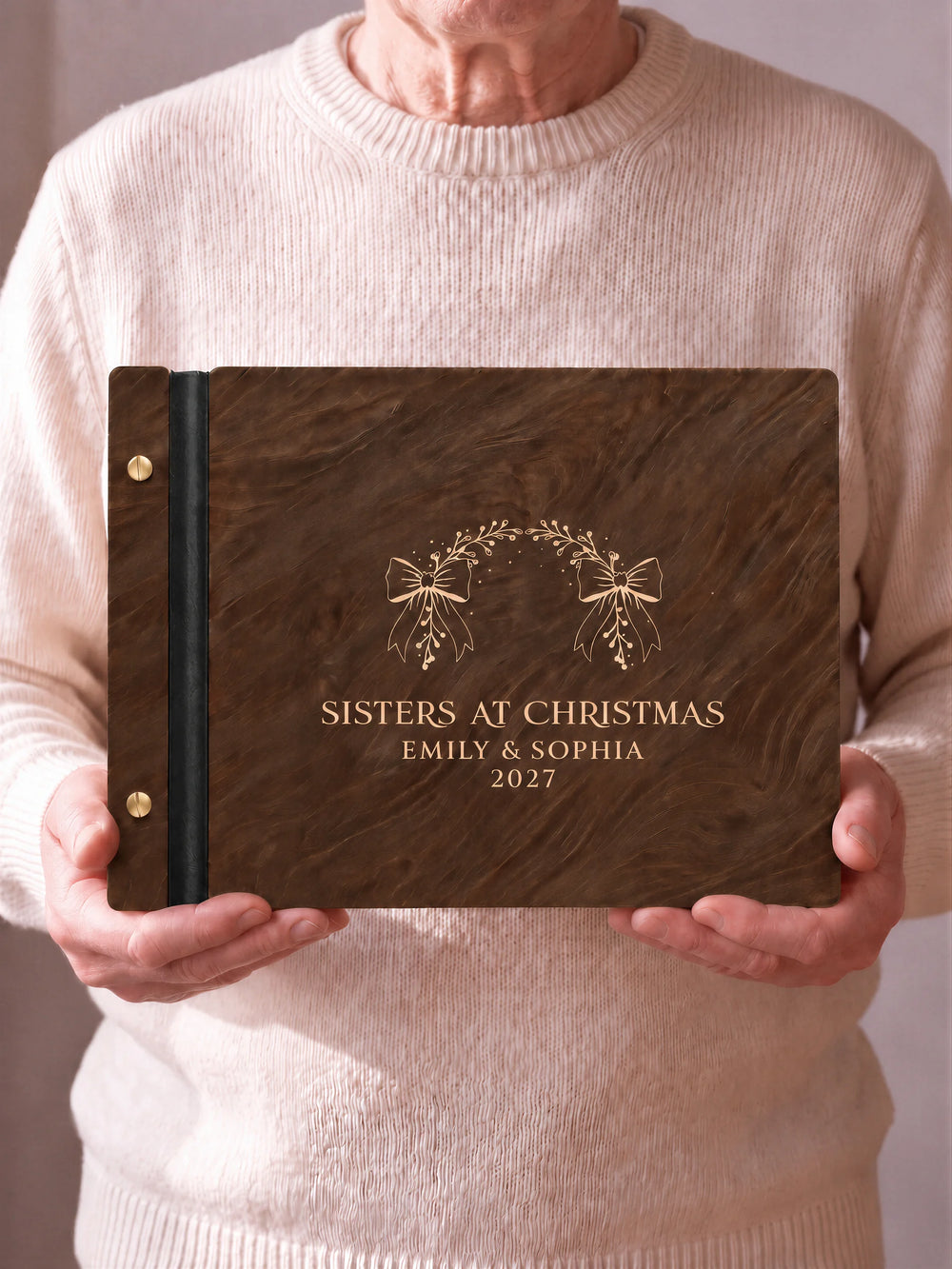 Two hands hold a Jacobean wooden photo album with the SISTERS AT CHRISTMAS cover facing the camera. Cream engraved artwork, the narrow binding and gold screws are visible.