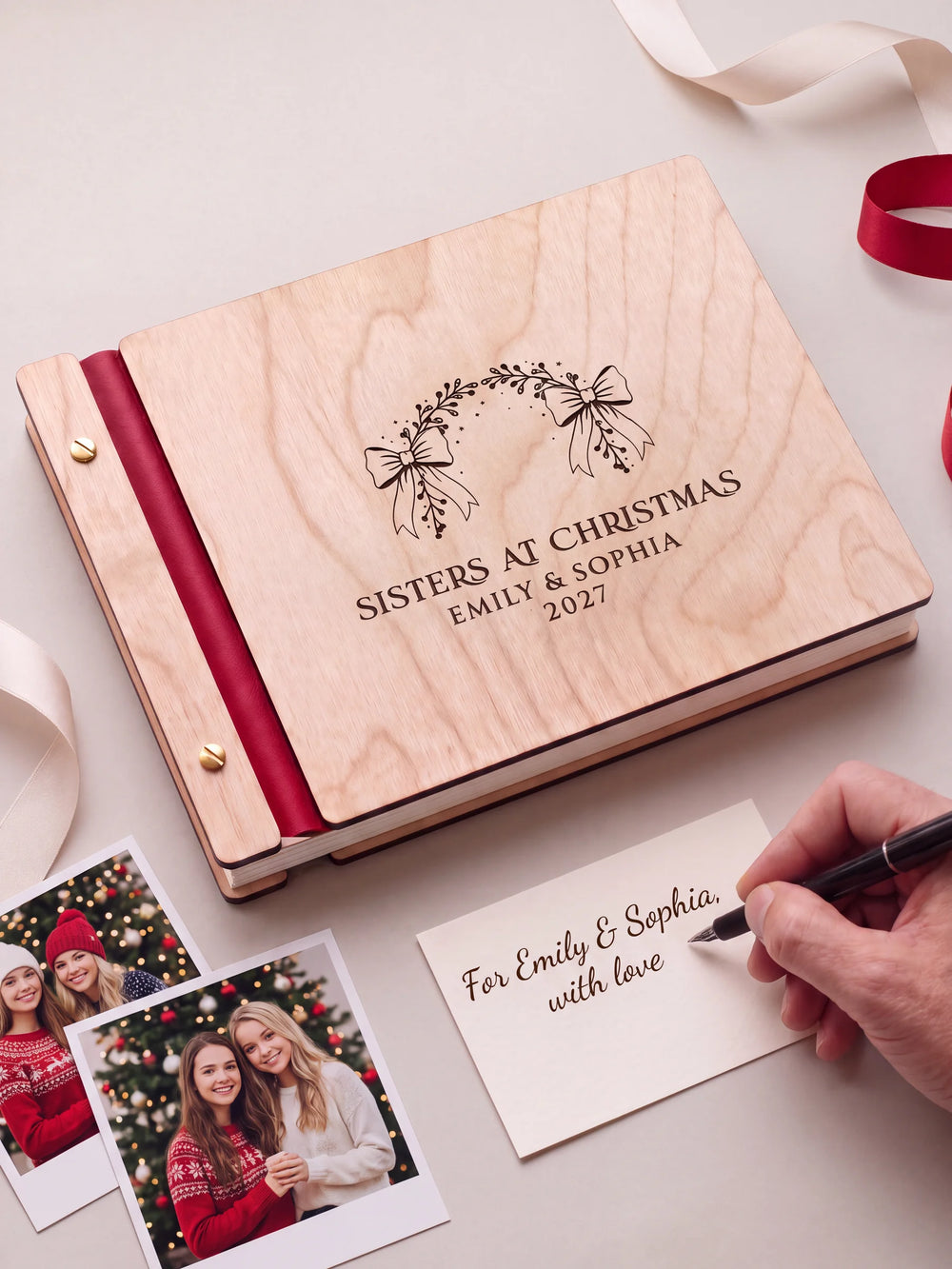 A Natural wooden album with the SISTERS AT CHRISTMAS engraving, cherry red binding and gold screws. A hand writes on a separate gift card beside the book.