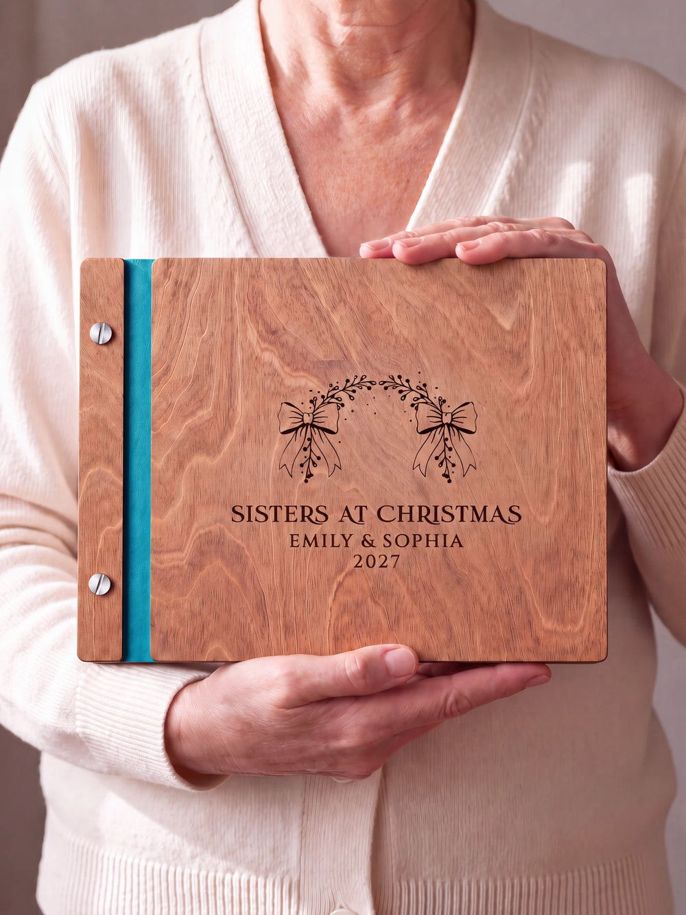 Two hands hold a Hazelnut wooden photo album. The SISTERS AT CHRISTMAS cover design has brown engraving, turquoise binding and silver screws.