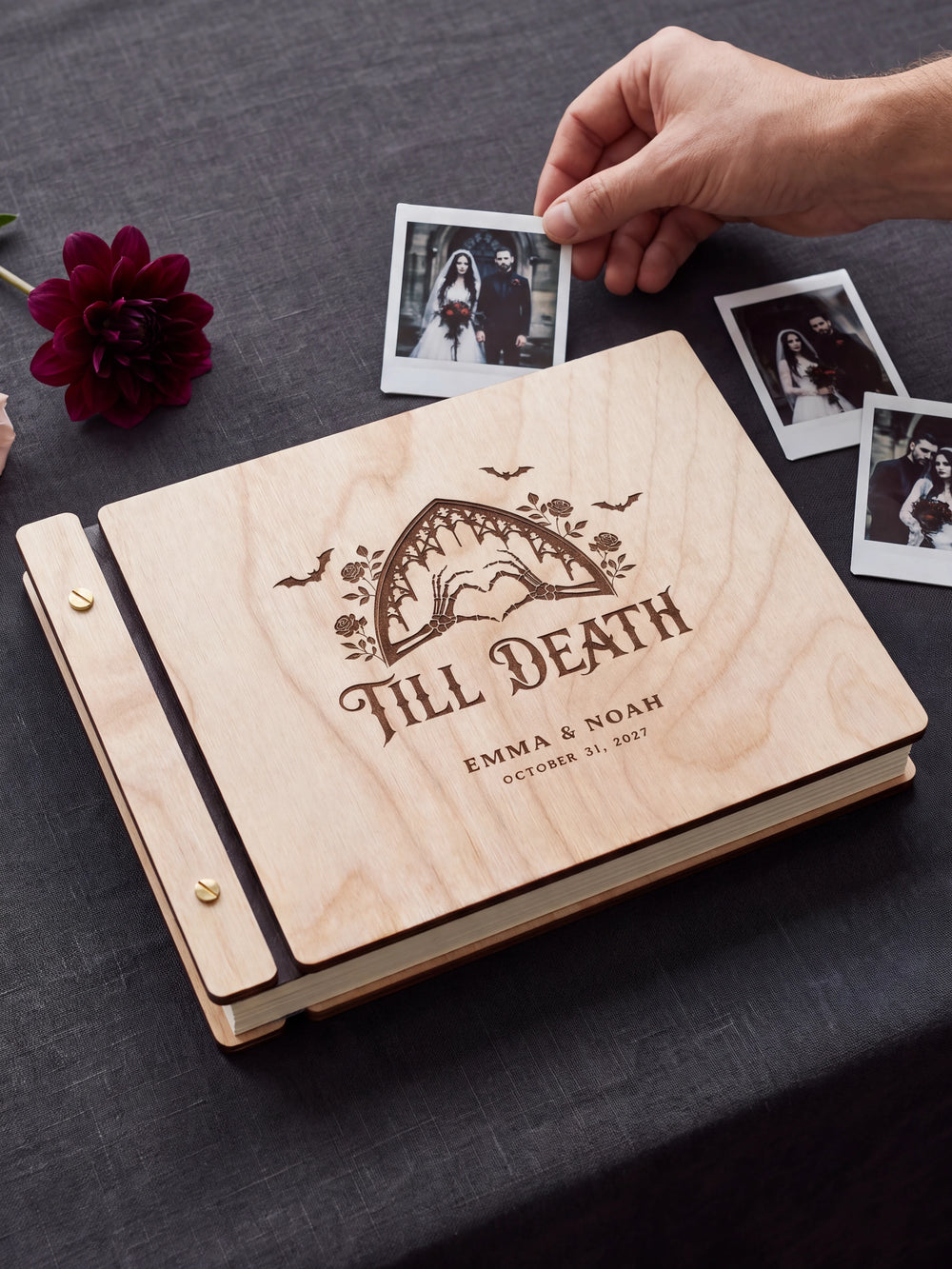Natural wood Till Death wedding photo guest book on a light table while a hand picks an instant photo to add; the engraved cover reads “TILL DEATH”, “EMMA & NOAH” and “OCTOBER 31, 2027”