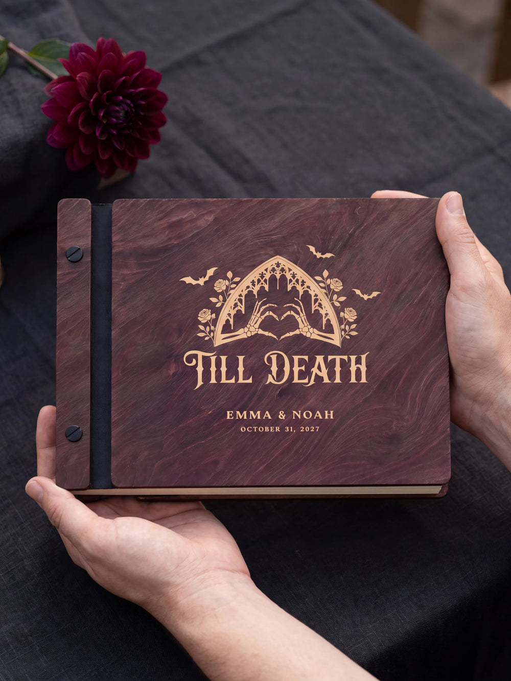 Two hands holding the Jacobean Till Death wedding photo guest book up to the camera, the cream engraving of a gothic arch with skeleton hands forming a heart and “TILL DEATH”, “EMMA & NOAH” and “OCTOBER 31, 2027” filling the cover