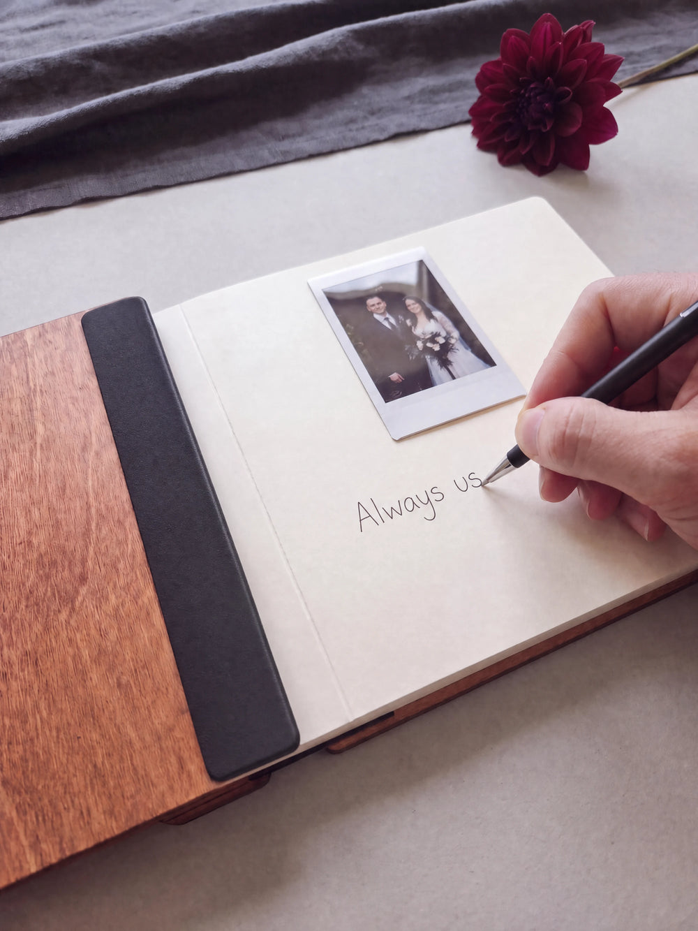 A hand writing a note in fine black pen beneath an instant photo on the cream page of the open Hazelnut Till Death wedding photo guest book