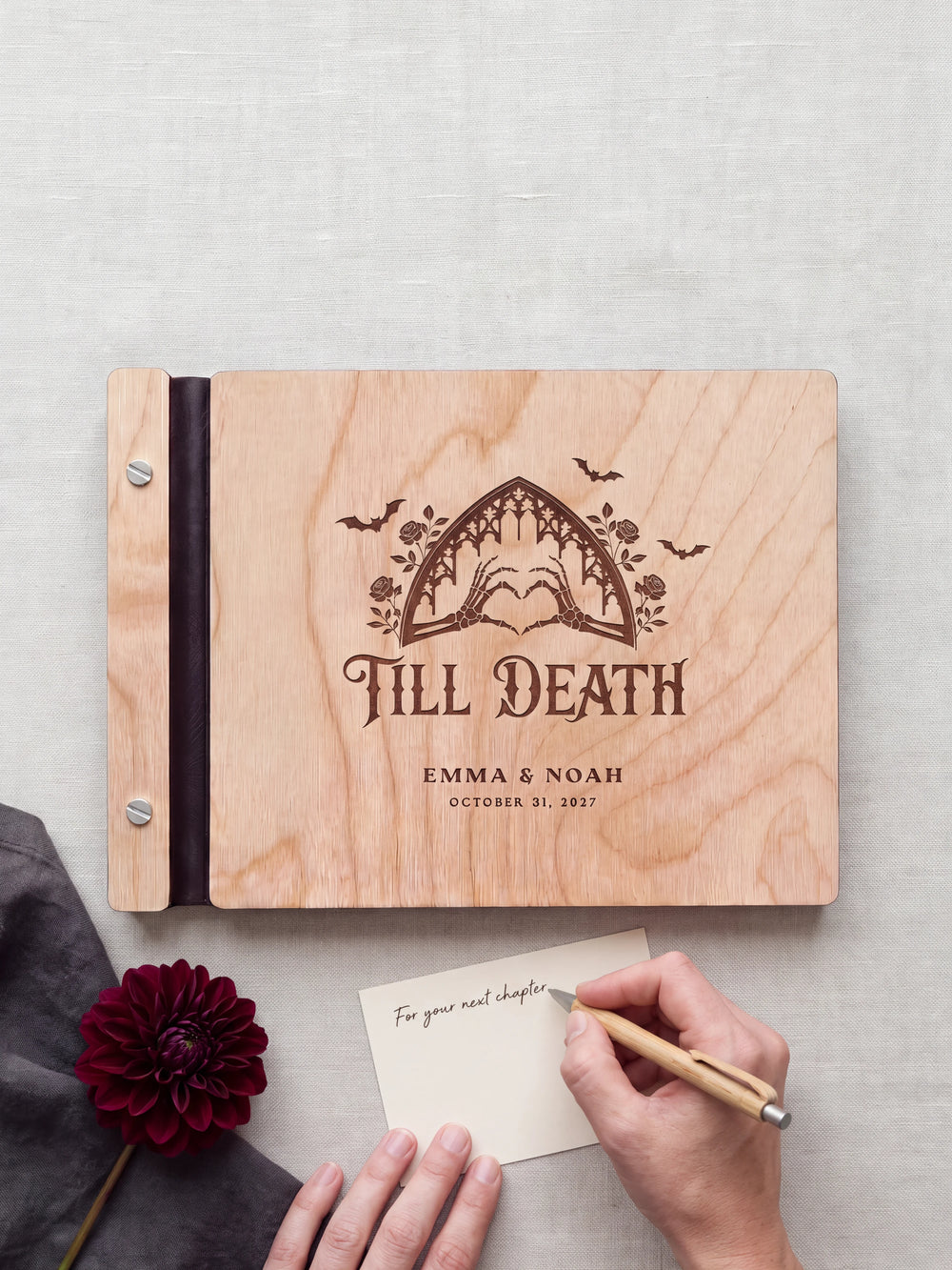 A hand writing a small gift card beside the closed Natural wood Till Death wedding photo guest book; the engraved cover and silver screws are visible