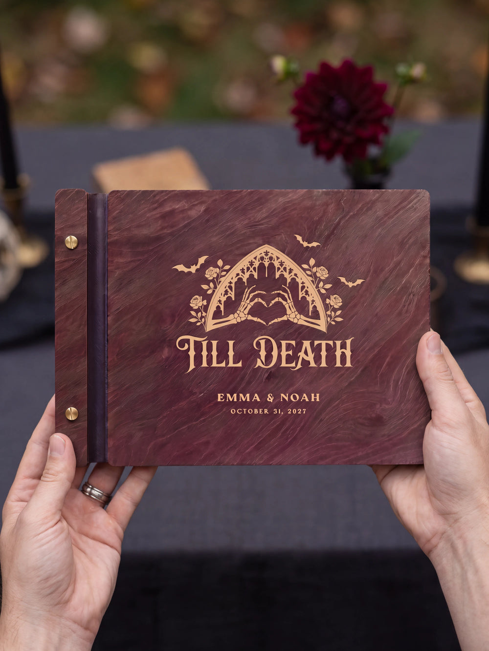 Two hands presenting the closed Jacobean Till Death wedding photo guest book with its cream engraving of a gothic arch with skeleton hands forming a heart, a single burgundy flower on the table beside them