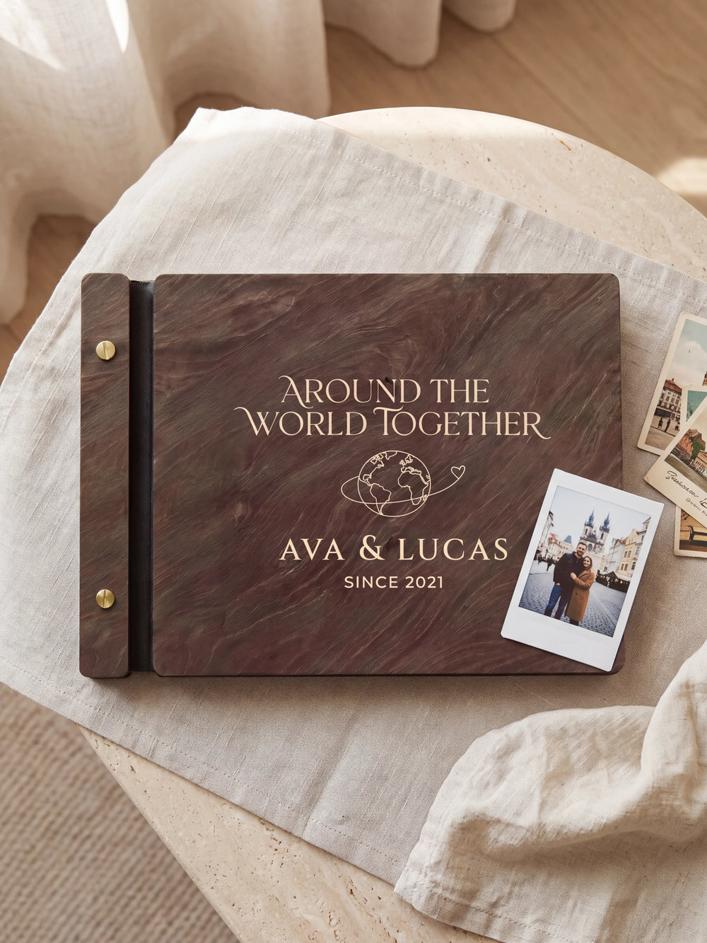 Dark brown Jacobean wooden album on a light surface, with cream engraving of a globe with a looping travel path and personalized cover wording. The full cover and left screw binding are visible.