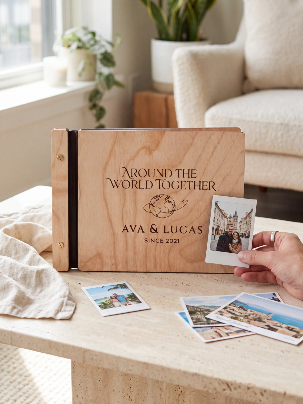 Natural wooden album with dark engraving of a globe with a looping travel path. A hand holds an instant photo beside the closed cover, with loose snapshots nearby.