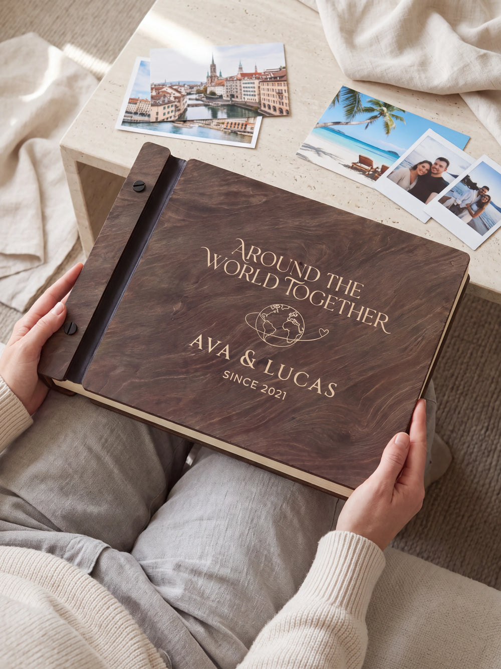 Hands hold a dark brown Jacobean wooden album close to the camera. Cream engraving shows a globe with a looping travel path, personalized wording and the black binding strip.