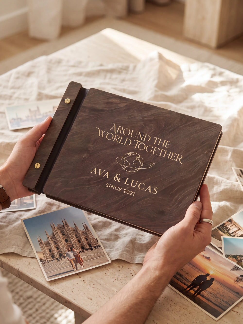 Two hands present a dark brown Jacobean wooden album, with the full cream-engraved globe with a looping travel path design and personalized cover wording facing the camera.