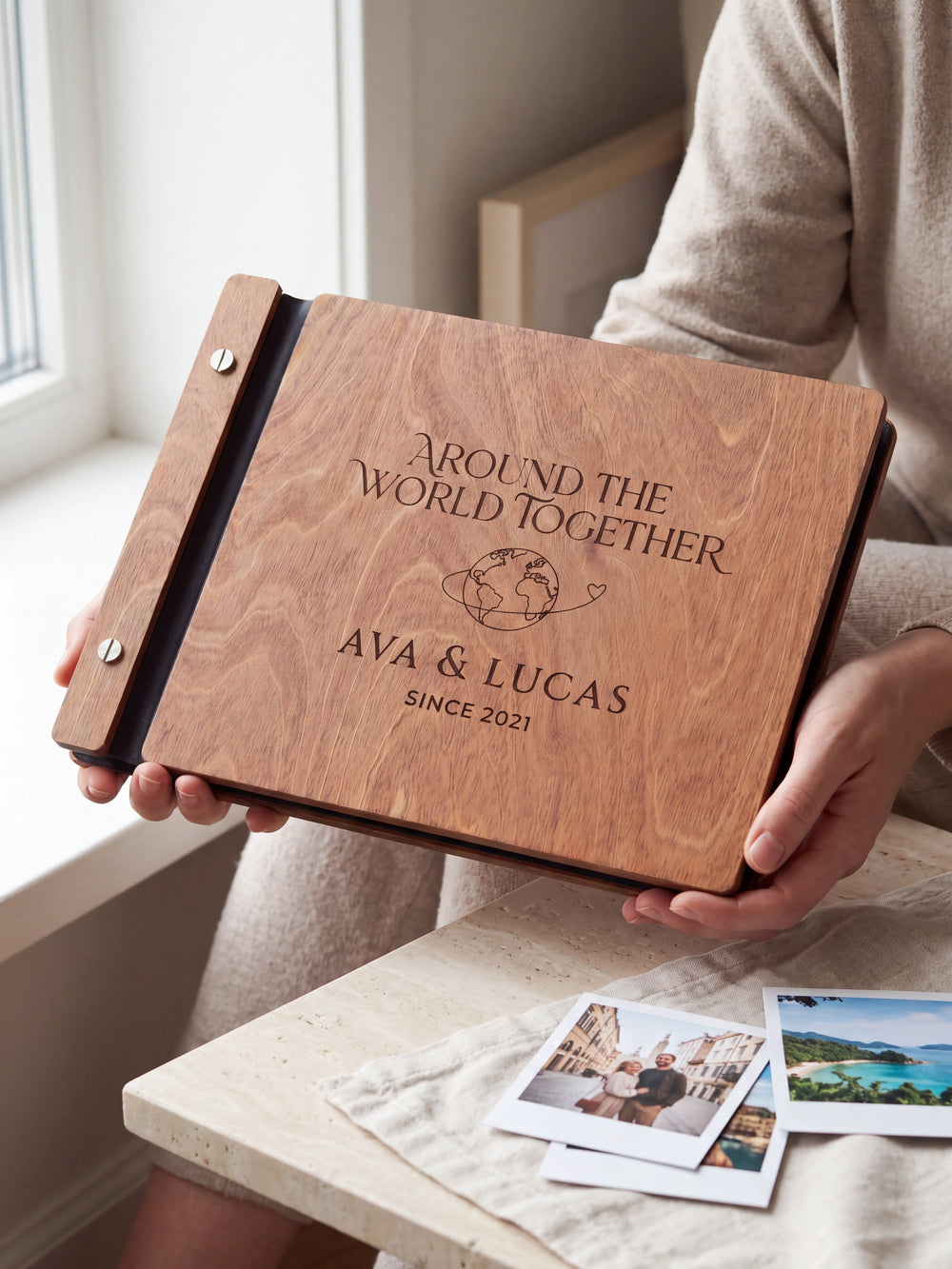 Hands hold the Hazelnut wooden album near a window. The full cover displays a globe with a looping travel path in dark engraving, with personalized lettering and a black binding strip.