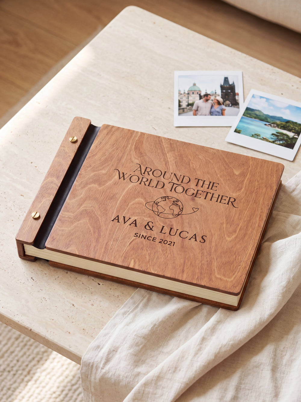 Hazelnut wooden album on a light tabletop with neutral linen. The full cover shows a globe with a looping travel path, personalized wording and the two-screw binding.