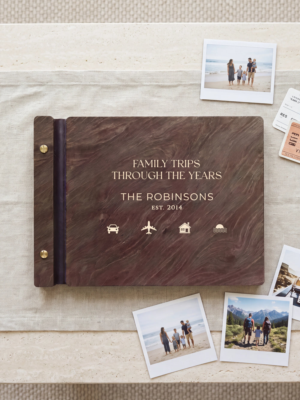 Dark brown Jacobean wooden album on a light surface, with cream engraving of small travel symbols and personalized cover wording. The full cover and left screw binding are visible.