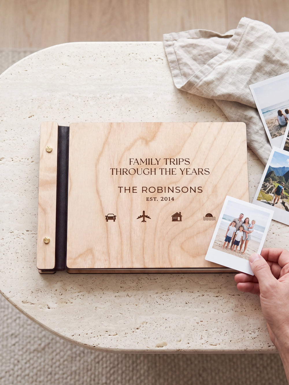 Natural wooden album with dark engraving of small travel symbols. A hand holds an instant photo beside the closed cover, with loose snapshots nearby.