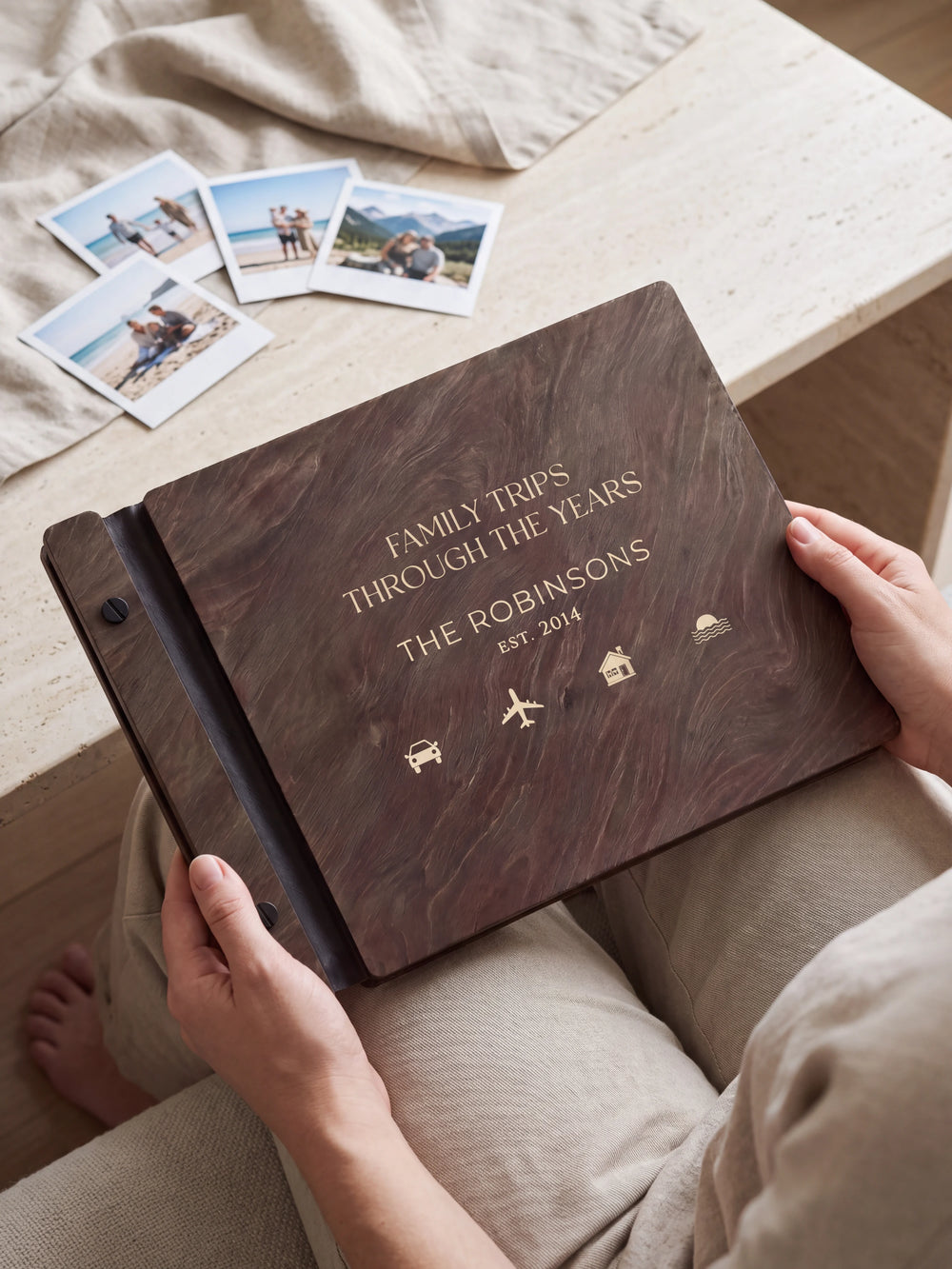 Hands hold a dark brown Jacobean wooden album close to the camera. Cream engraving shows small travel symbols, personalized wording and the black binding strip.