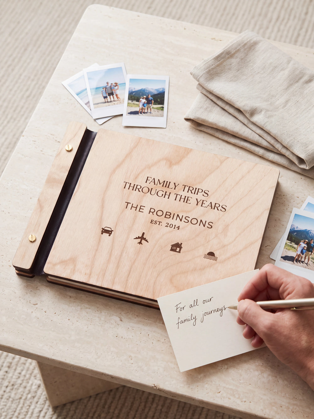 A Natural wooden album showing small travel symbols rests beside a separate gift card. A hand writes 
