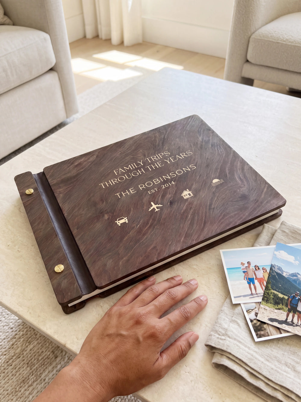 A hand rests beside the closed dark brown Jacobean album on a table. Cream engraving and personalized wording are visible, with travel photos nearby.