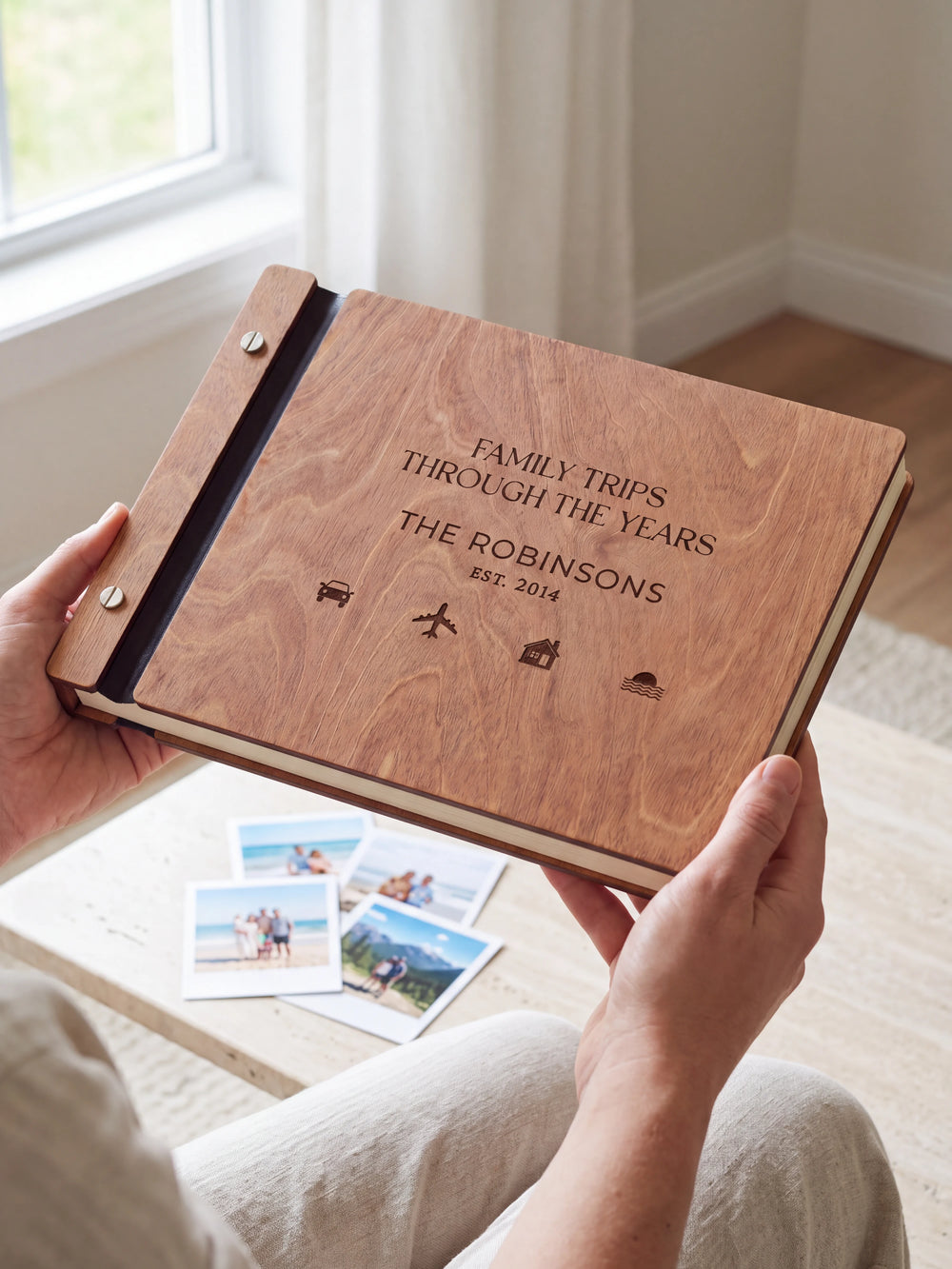 Hands hold the Hazelnut wooden album near a window. The full cover displays small travel symbols in dark engraving, with personalized lettering and a black binding strip.