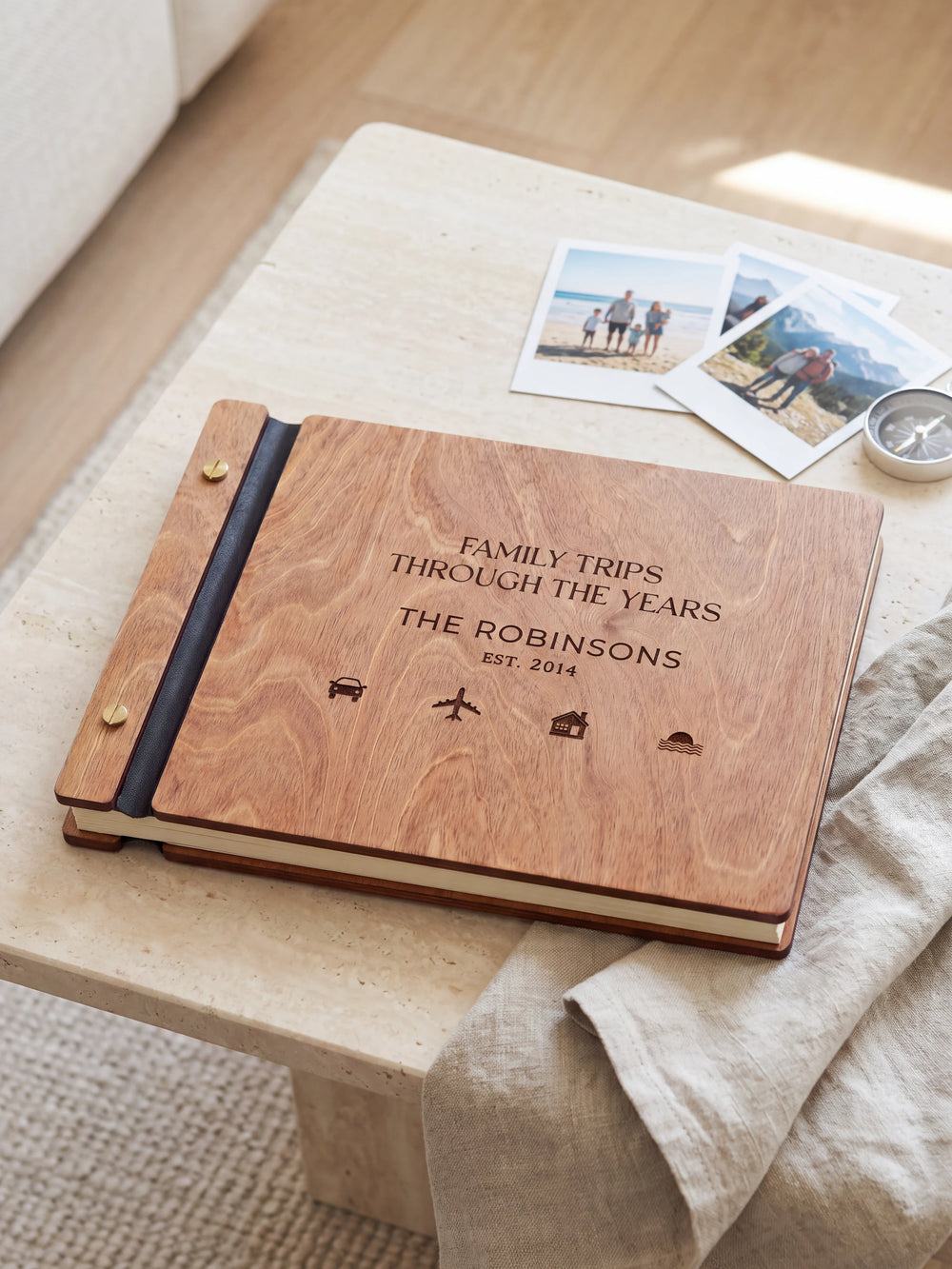 Hazelnut wooden album on a light tabletop with neutral linen. The full cover shows small travel symbols, personalized wording and the two-screw binding.