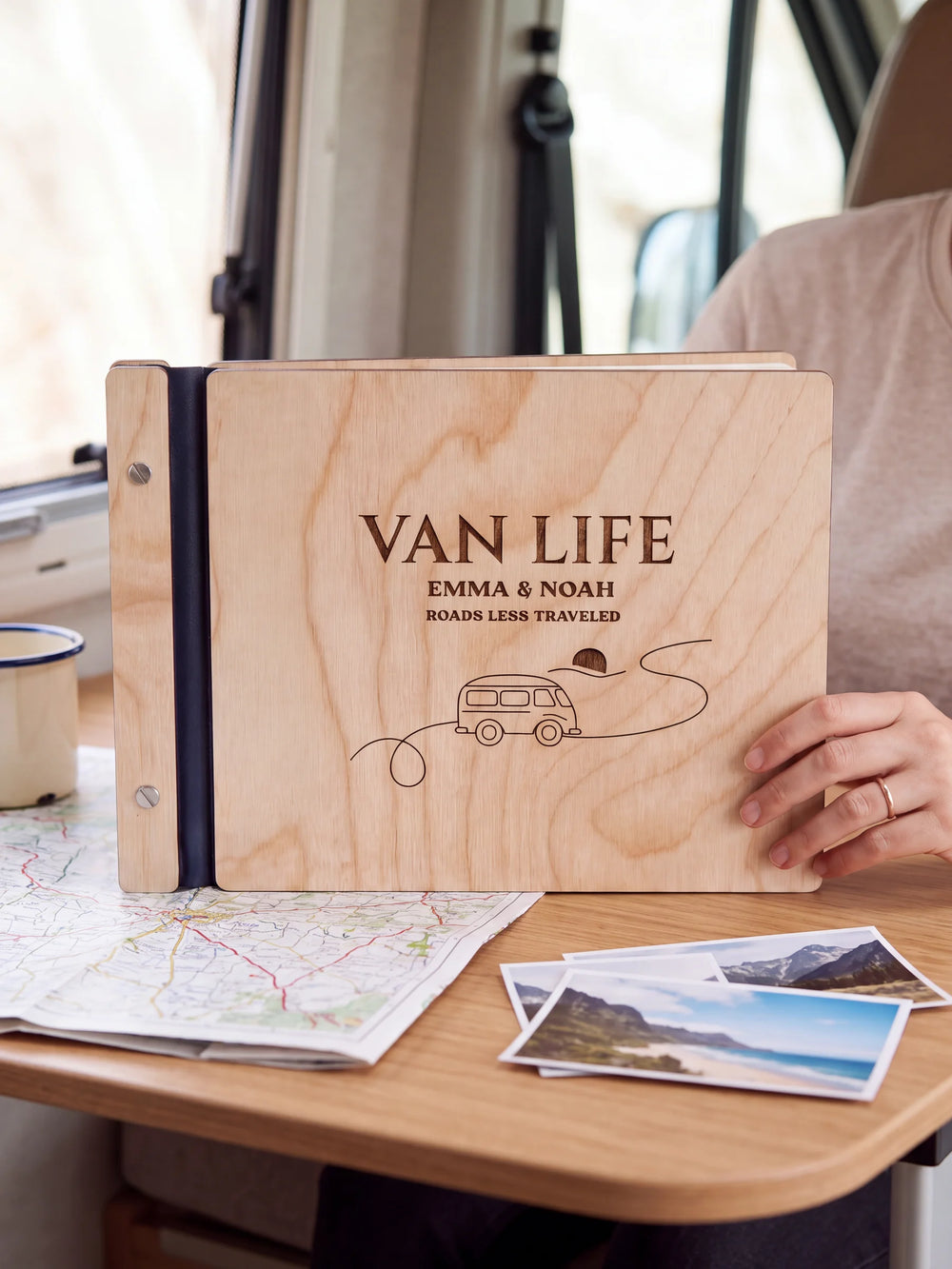 Natural wooden album with brown laser engraving of camper van, winding road and sun, shown in a close lifestyle view with navy blue binding and silver screws. The cover reads VAN LIFE.