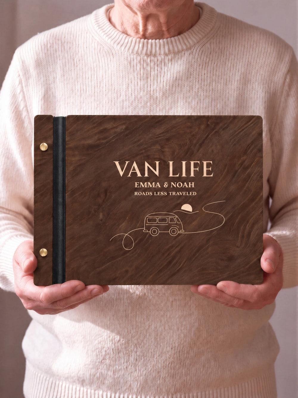 Two hands hold a Jacobean wooden photo album with the VAN LIFE cover facing the camera. Cream engraved artwork, the narrow binding and gold screws are visible.