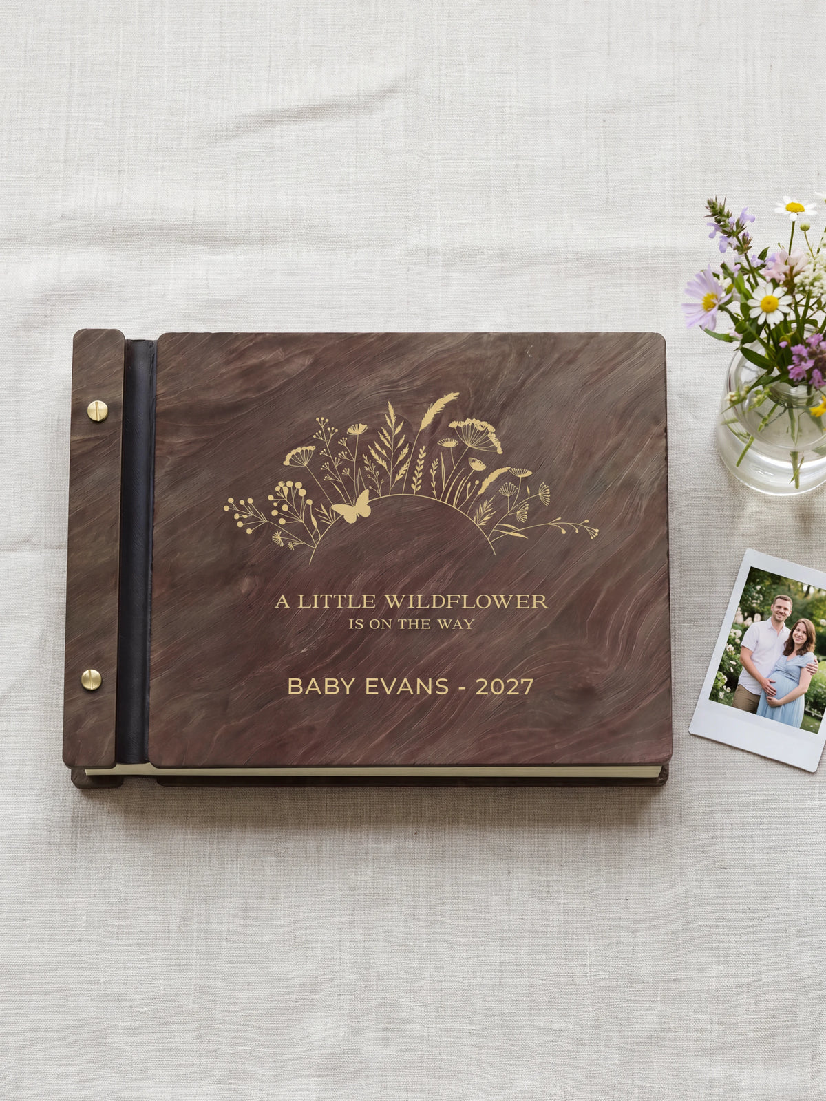 Jacobean wood wildflower baby shower photo guest book lying flat on ivory linen, the cover engraved with “A LITTLE WILDFLOWER IS ON THE WAY” and “BABY EVANS - 2027”; two gold-tone screws along the black vegan leather spine
