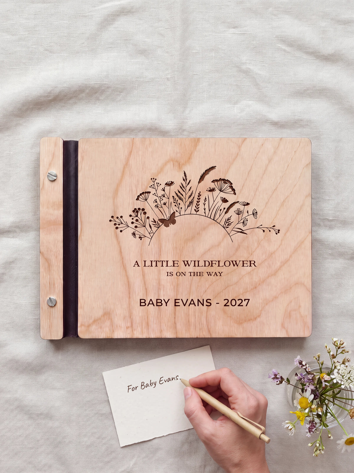 A hand writing a small gift card beside the closed Natural wood wildflower baby shower photo guest book; the engraved cover and silver screws are visible