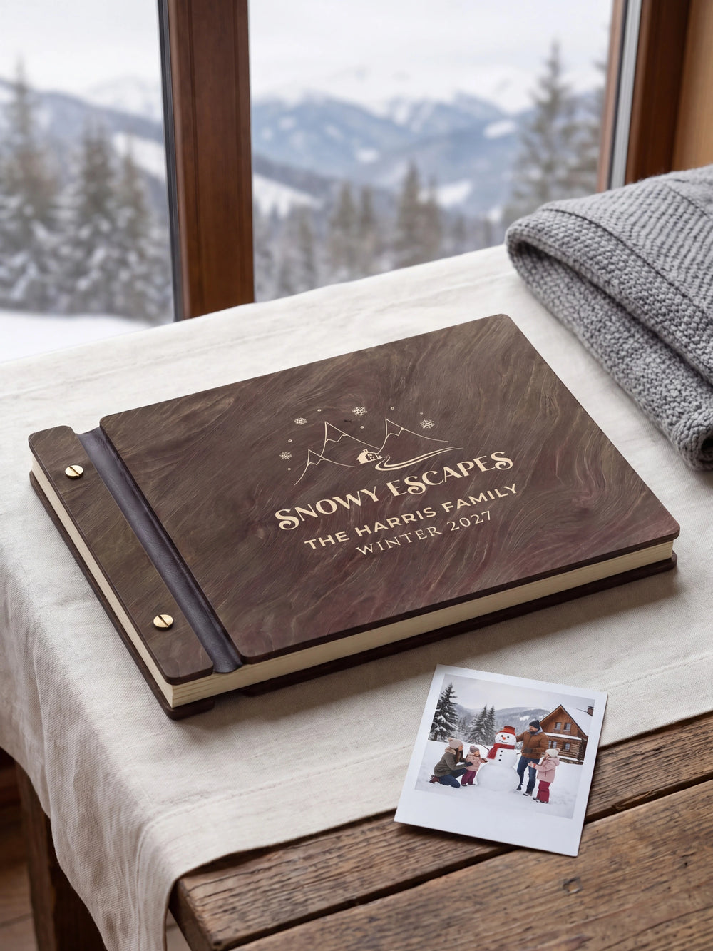 Dark brown Jacobean wooden album on a light surface, with cream engraving of snowy mountains, a cabin and snowflakes and personalized cover wording. The full cover and left screw binding are visible.