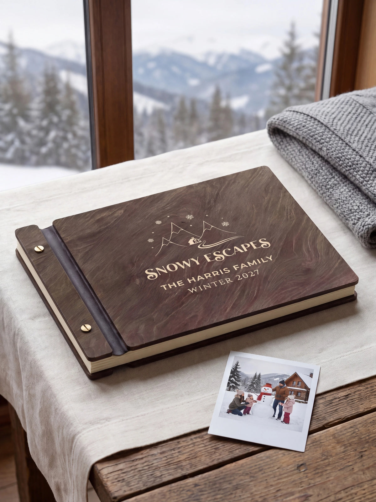 Dark brown Jacobean wooden album on a light surface, with cream engraving of snowy mountains, a cabin and snowflakes and personalized cover wording. The full cover and left screw binding are visible.