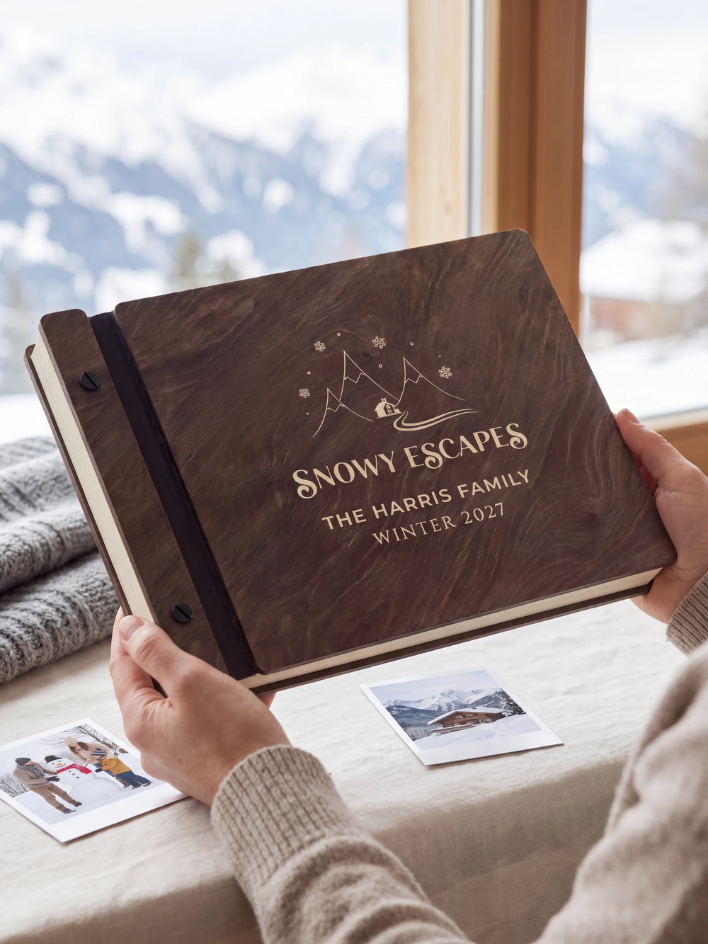 Hands hold a dark brown Jacobean wooden album close to the camera. Cream engraving shows snowy mountains, a cabin and snowflakes, personalized wording and the black binding strip.